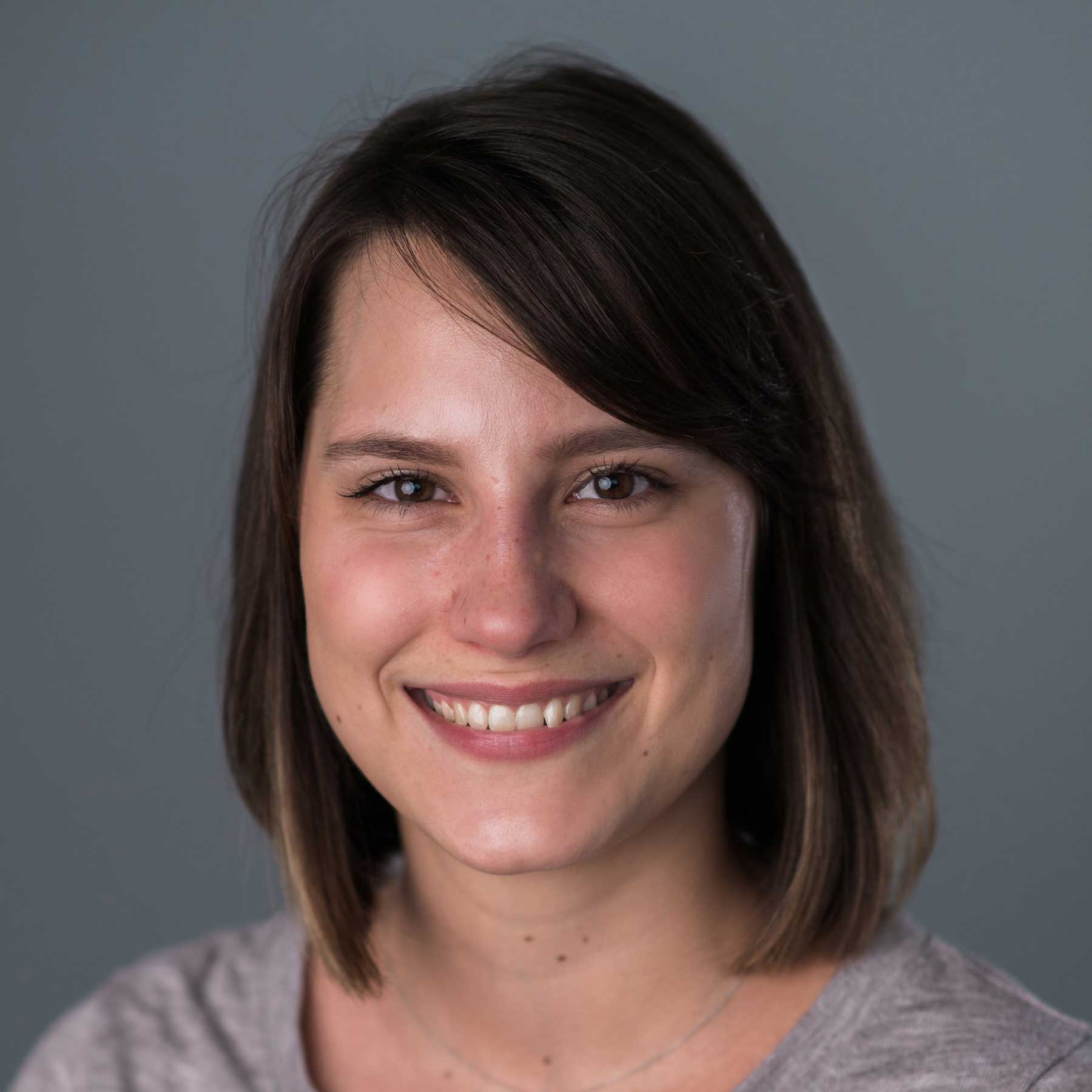 A woman with straight brown hair, wearing a grey shirt, smiles at the camera against a plain grey background.