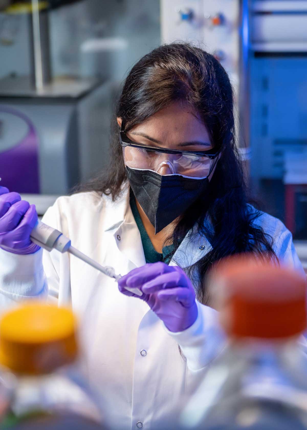 A scientist wearing a lab coat, safety goggles, mask, and gloves uses a pipette to transfer liquid in a laboratory setting with equipment visible in the background.