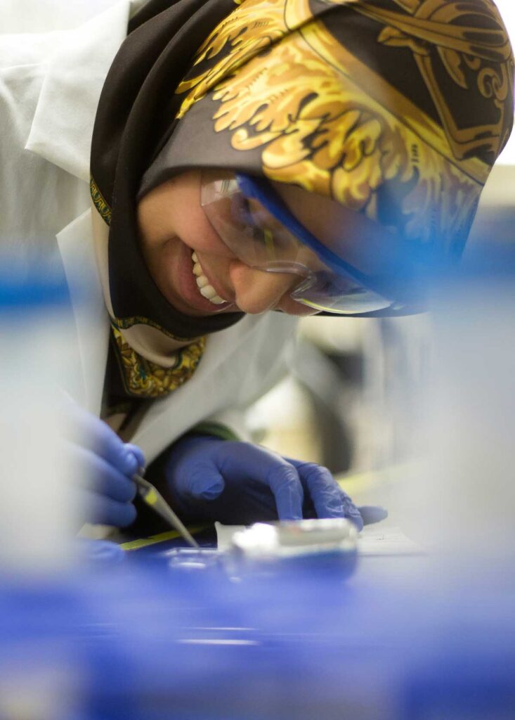 A scientist in a patterned headscarf and safety glasses works at a lab bench, using a tool with gloved hands.