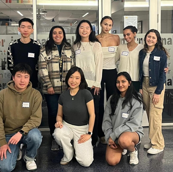 A group of nine people poses indoors, most wearing nametags and casual clothing, standing and kneeling in two rows, with a glass wall and office setting in the background.