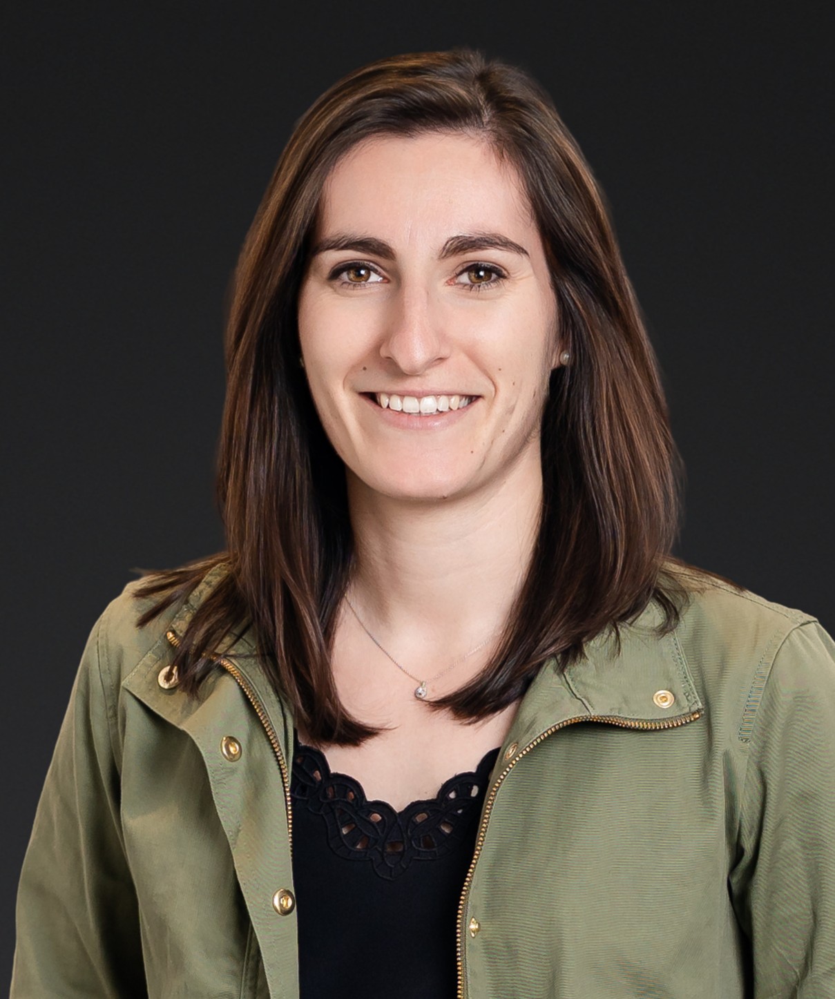 A woman with straight brown hair wearing a green jacket over a black top, smiling at the camera against a plain dark background.